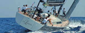 Group of people on a white luxury sailing yacht leaning to one side as it slices through blue ocean waves, with crew on deck attending to controls and sails.